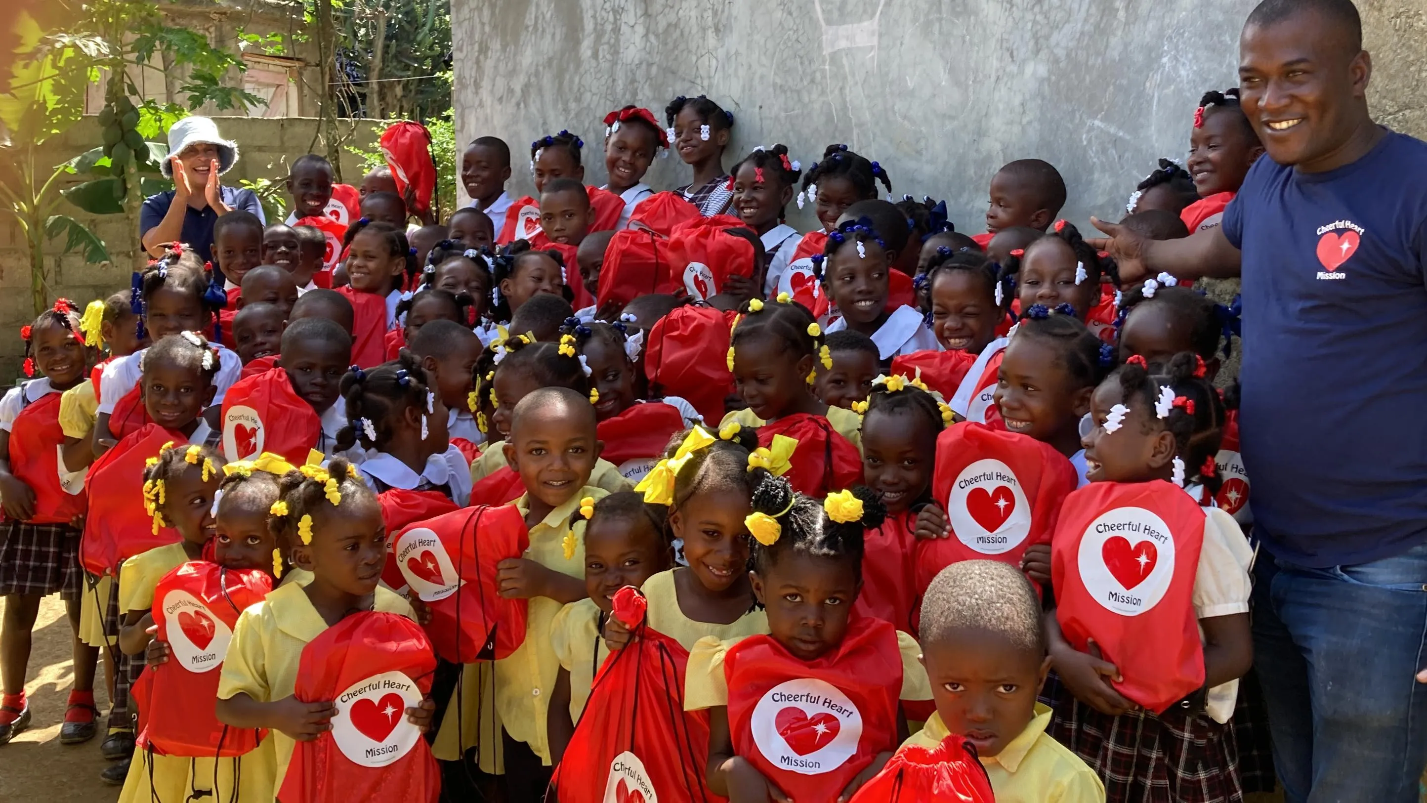 A large group of smiling schoolchildren hold red Cheerful Heart Mission bags alongside a staff member