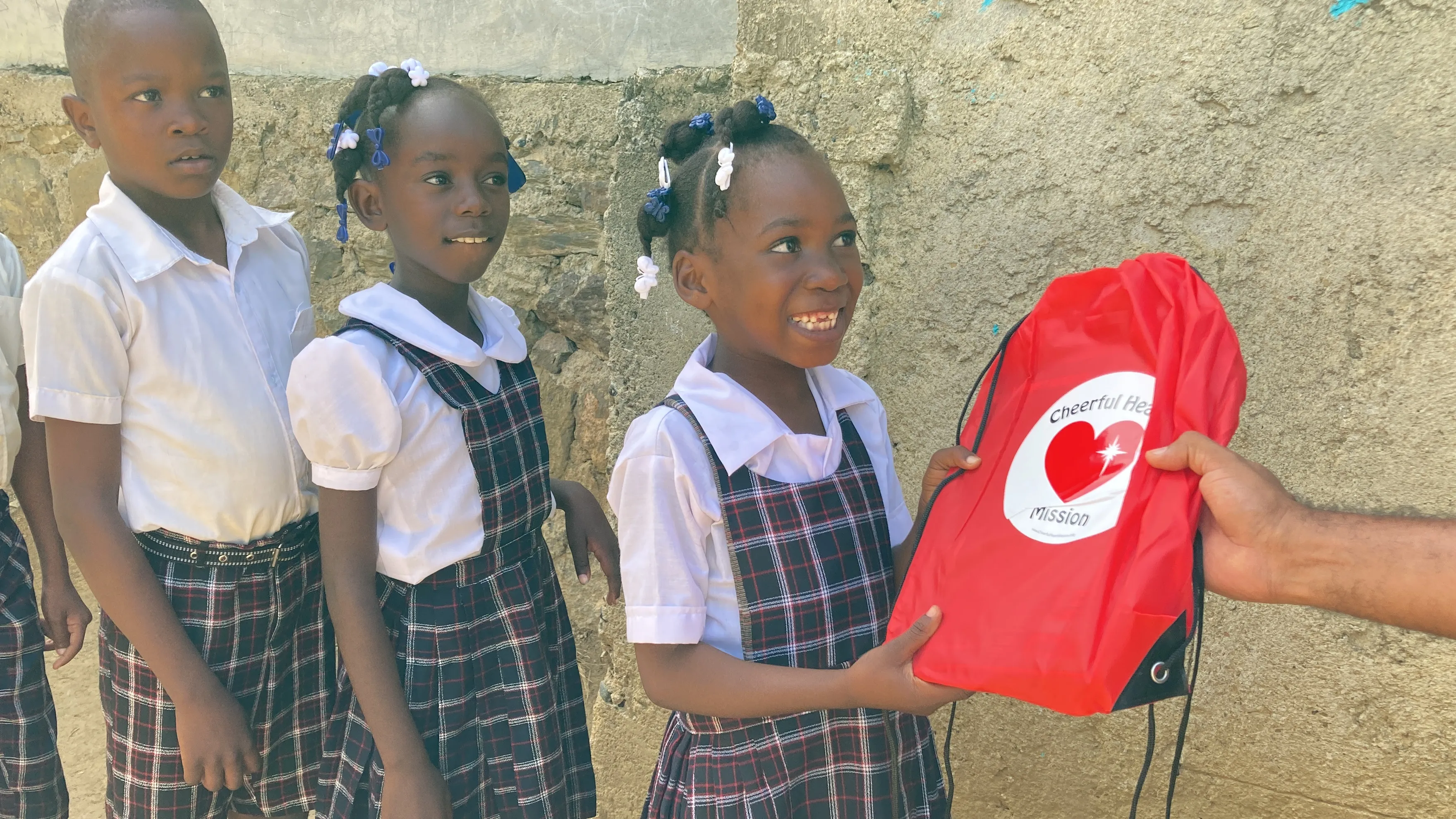 A smiling girl in a plaid school uniform receives a red Cheerful Heart Mission backpack